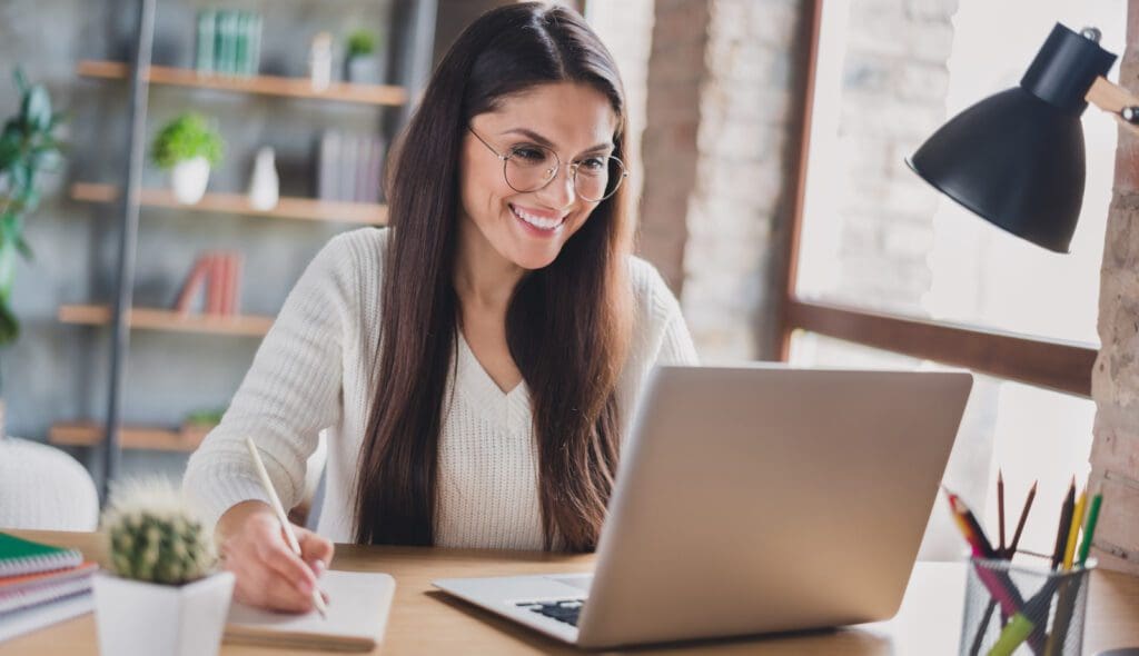 Photo of lady sit desk look screen laptop hold pen write notepad wear spectacles white sweater in living room home indoors Author working on author website