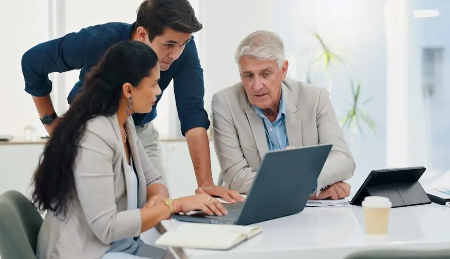Laptop, teamwork and business people in office, conversation or planning for property development. Employees, coworking or group in meeting, pc or internet for real estate with discussion or advice A man working on digital estate planning with two helpers.