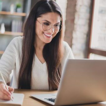 Photo of lady sit desk look screen laptop hold pen write notepad wear spectacles white sweater in living room home indoors Author working on author website